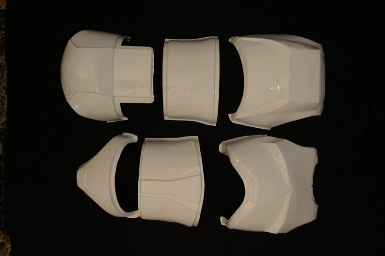 May include: White, segmented armor pieces arranged on a dark surface. The set includes various curved and angular components, likely for a costume or prop. The pieces have a glossy finish and appear to be made of a hard material.