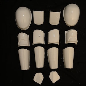 May include: A collection of white, glossy plastic armor pieces, including helmet sections, shoulder pads, and shin guards, arranged on a dark surface. The pieces vary in size and shape, suggesting a full body armor set.