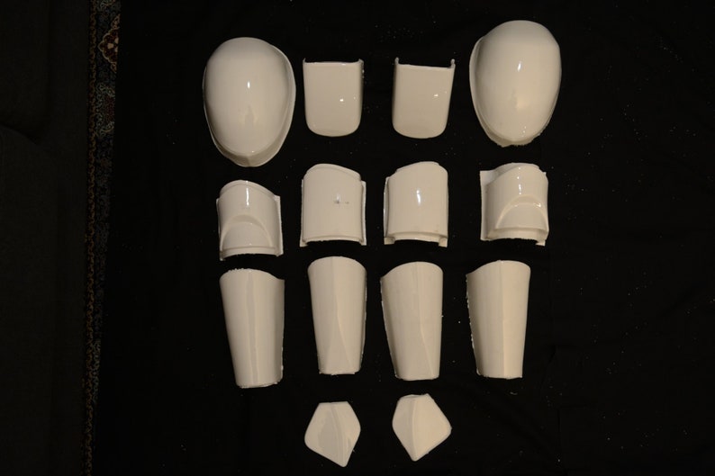 May include: A collection of white, glossy plastic armor pieces, including helmet sections, shoulder pads, and shin guards, arranged on a dark surface. The pieces vary in size and shape, suggesting a full body armor set.
