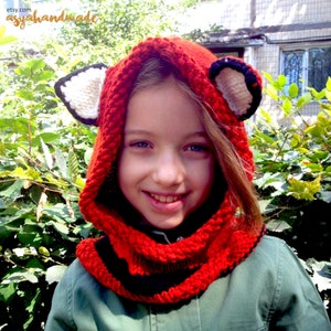 May include: A red knitted fox hat with black and white stripes and fox ears. The hat is worn by a child with long brown hair.