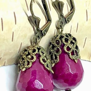 May include: A pair of drop earrings featuring faceted, teardrop-shaped magenta beads. Each bead is topped with an ornate, antique bronze-colored metal filigree cap and finished with a small bronze-colored bead at the bottom, attached to leverback hooks.