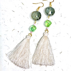 May include: A pair of earrings with white tassel charms. The earrings feature green and clear faceted beads and gold-toned metal.