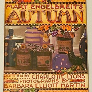 May include: A decorative book cover titled "Mary Engelbreit's Autumn" with illustrations of jars of preserves, a black bird, and autumn foliage. The book cover includes the text "Written by Charlotte Lyons" and "Photographs by Barbara Elliott Martin."