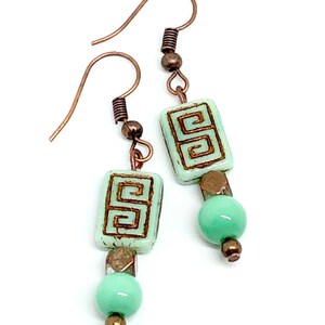 May include: A pair of dangle earrings with a copper wire hook, a light green glass bead, and a light green square bead with a brown geometric design.