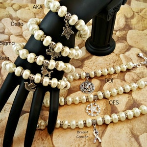 May include: A set of six pearl bracelets with silver charms. The charms include a star, a bird, a dove, a compass, a star with rhinestones, and a pink ribbon. The bracelets are labeled with the Greek letters AKA, Delta, Sigma, Zeta, DOI, and OES. The bracelets are on a black hand model against a background of brown stones.