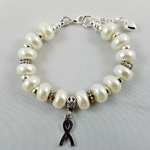 May include: A white pearl bracelet with a silver charm in the shape of a ribbon. The bracelet has a silver clasp and a small silver heart charm.