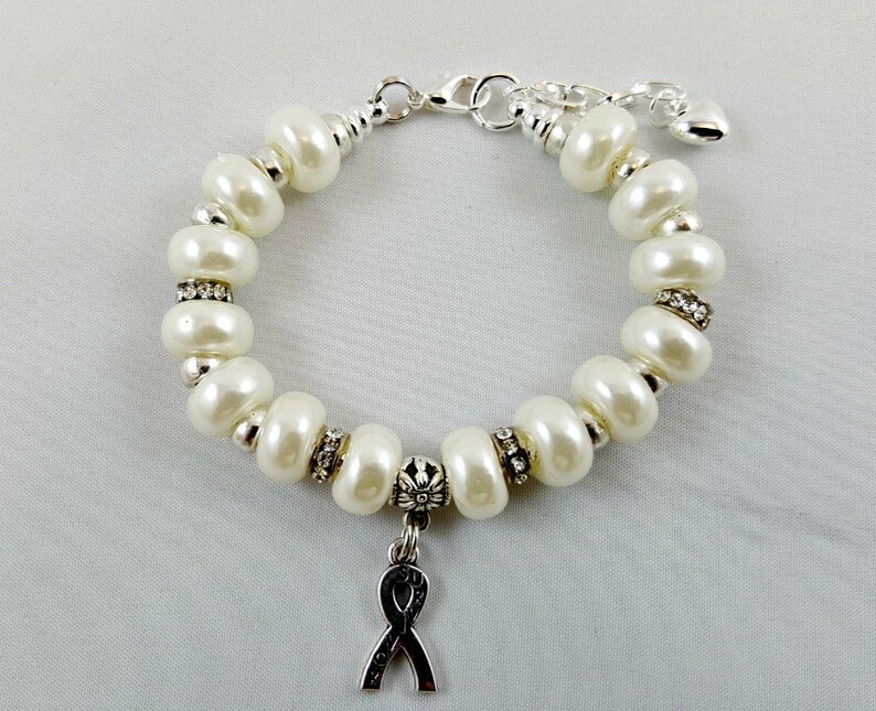 May include: A white pearl bracelet with a silver charm in the shape of a ribbon. The bracelet has a silver clasp and a small silver heart charm.
