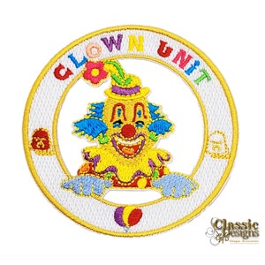 Puede incluir: Un parche redondo bordado blanco con una cara de payaso amarilla en el centro. El payaso lleva un sombrero morado, un cuello azul y blanco y una camisa a rayas rojas y blancas. Las palabras "Clown Unit" están escritas en un círculo alrededor del payaso.