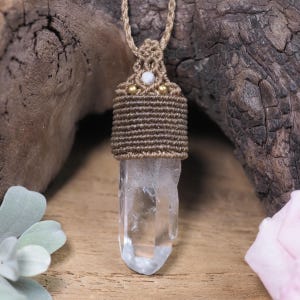 May include: A clear quartz crystal pendant with a macrame cord. The pendant is wrapped in brown macrame and features a small white bead and gold-coloured accents. The cord is a natural, light brown colour.