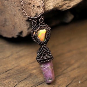 May include: A handmade macramé necklace with an iridescent opal stone at the centre and a lower, elongated, purple stone. The necklace is made with brown cord, and the stones are set in intricate macramé knots. The background is a wooden surface.