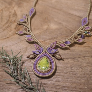 May include: A macrame necklace featuring a central teardrop-shaped green stone surrounded by concentric rings of purple and tan cord. The necklace band is adorned with symmetrical leaf-shaped patterns in purple and tan, with small accent beads.