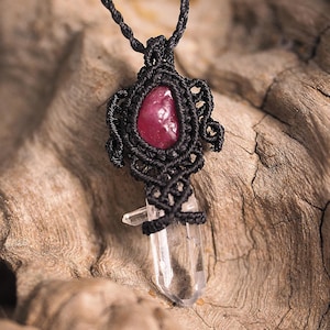 May include: A pendant necklace featuring a raw, deep pink gemstone set in intricate black macrame cord work, positioned above a clear, pointed quartz crystal point that is also secured within the same black knotted cord design.