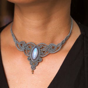 May include: A grey macramé choker necklace featuring an oval-shaped iridescent moonstone cabochon at the centre. The intricate knotted cord design includes small metallic gold-toned beads integrated into the pattern, forming a symmetrical shape.