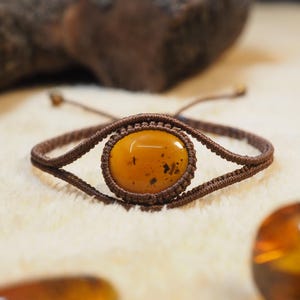 May include: A macrame bracelet with a central oval amber stone. The amber is a warm, golden-yellow colour with dark inclusions. The bracelet is made with brown macrame cord and has adjustable sliding knots.