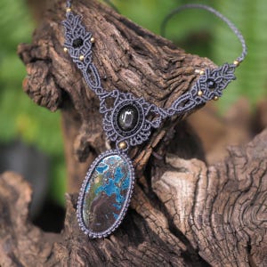 Pode incluir: Um colar de macramé artesanal com um grande pingente oval com pedra azul e castanha. O colar tem um design de macramé cinzento com detalhes em preto e dourado. O colar é exibido em um pedaço de madeira.