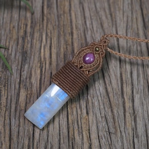 May include: A pendant necklace featuring a rectangular, translucent white moonstone with blue iridescence, topped with a brown macrame cord wrap and a small, round, dark pink gemstone set in a decorative, woven brown cord pattern.