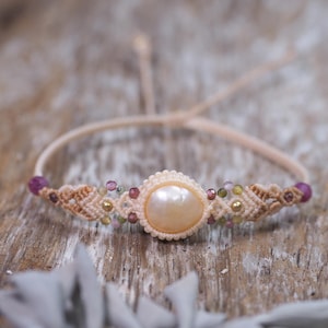 May include: A macrame bracelet featuring a central round peach-coloured pearl surrounded by small, multi-coloured faceted gemstone beads and gold-toned metal accents. The band is made of light beige cord with adjustable sliding knot closures.