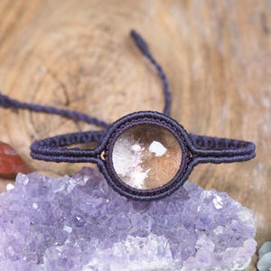 May include: A bracelet featuring a circular, translucent, amber-toned gemstone encased in a dark purple macrame cord setting. The cord is woven into a thin, adjustable band that extends outwards from the central stone setting on both sides.