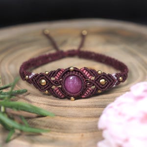 May include: A macrame bracelet featuring a central round deep pink gemstone surrounded by dark maroon and light pink knotted cord. The design includes small metallic gold-toned beads integrated into the woven pattern and adjustable cord ends.