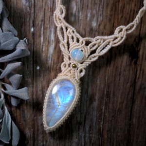 May include: A macrame necklace featuring a large, pear-shaped moonstone pendant with a blue iridescent sheen, accented by a smaller round moonstone and a tiny gold-toned bead, all set within intricate, cream-colored knotted cord work.