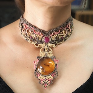 May include: A handcrafted macrame necklace featuring a large amber pendant. The necklace has a brown, pink, and beige color scheme with a central oval amber stone and smaller red gemstones. The intricate design is visible in the close-up shot.