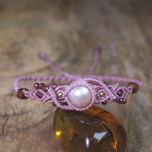 May include: A macrame bracelet featuring a central iridescent white pearl, small dark red faceted garnet beads, and tiny gold-toned metal spheres. The bracelet is constructed from light pink cord with an adjustable sliding knot closure system.
