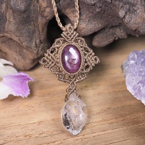 May include: A handcrafted macrame necklace featuring a central oval-shaped, pink gemstone pendant. The pendant is encased in intricate macrame work, with a clear crystal pendant hanging below. The necklace is displayed on a wooden surface.
