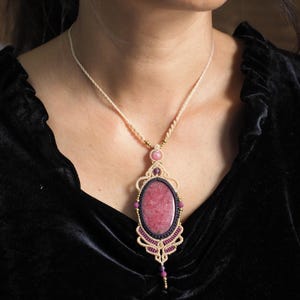 May include: A macrame pendant necklace featuring a large oval pink stone set in intricate beige and dark purple cord work. The design includes small round pink and purple beads, metallic accents, and a braided beige cord necklace chain.