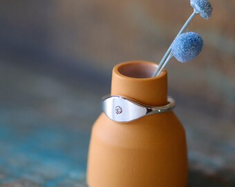 May include: A silver ring with a single diamond set in an oval-shaped band. The ring is displayed on a small, orange ceramic vase.