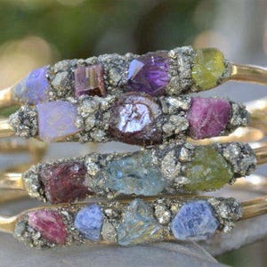 May include: Four gold bangles stacked, each adorned with a cluster of colourful gemstones. The gemstones include purple, pink, blue, and green hues, set in a textured, silver-toned metal. The bangles have a simple, elegant design.