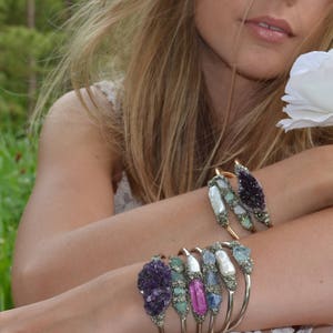 May include: A collection of gold-toned bracelets adorned with various gemstones, including amethyst, pearls, and opals. The bracelets are displayed on a person's arm, with a soft focus background of greenery and a white flower.