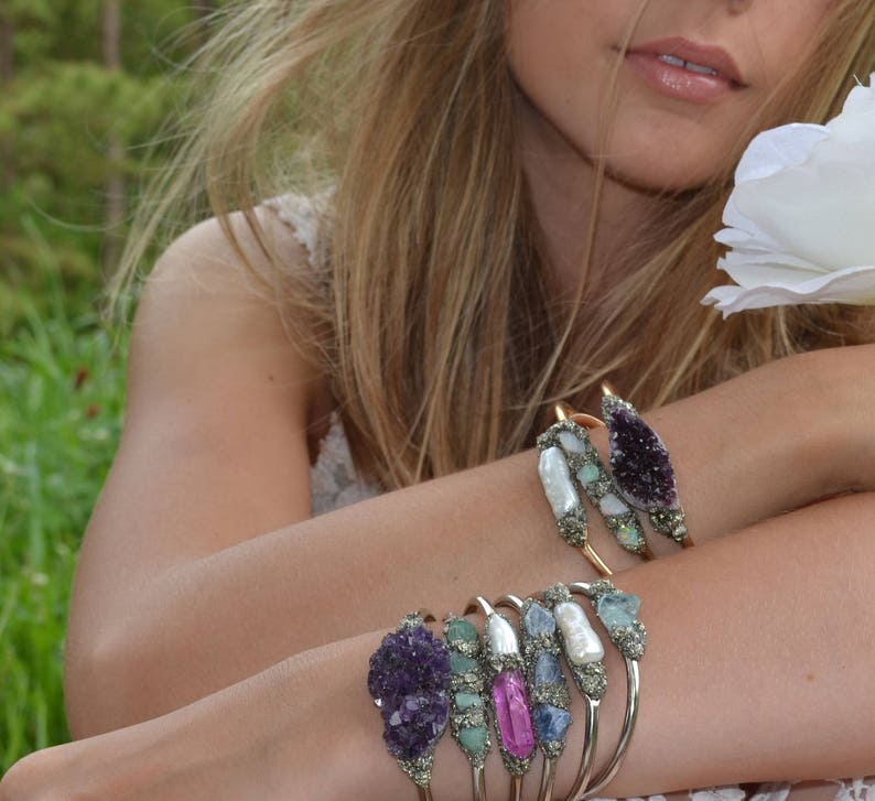 May include: A collection of gold-toned bracelets adorned with various gemstones, including amethyst, pearls, and opals. The bracelets are displayed on a person's arm, with a soft focus background of greenery and a white flower.