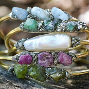 May include: Stacked gold-tone bracelets, each adorned with a unique arrangement of raw gemstones. The bracelets feature a variety of colors, including blue, green, pink, and white stones, set against a textured, silver-toned base.