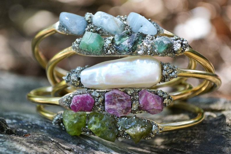 May include: Stacked gold-tone bracelets, each adorned with a unique arrangement of raw gemstones. The bracelets feature a variety of colors, including blue, green, pink, and white stones, set against a textured, silver-toned base.
