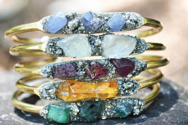 May include: Stack of gold-toned metal bracelets, each featuring a different colored gemstone. The gemstones include blue, white, red, orange, and green. The gemstones are set in a silver-colored matrix.