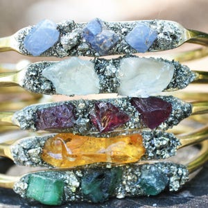 May include: A set of five gold bracelets with different colored gemstones and pyrite. The gemstones are blue, white, red, orange, and green.