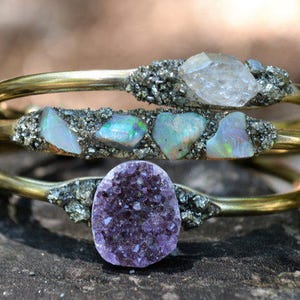 May include: Three gold-toned metal bracelets with natural stone accents. The top bracelet features a clear crystal, the middle one has opals, and the bottom one showcases a purple amethyst. The stones are set in a textured, silver-colored material.