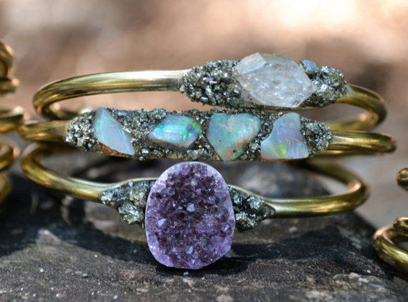 May include: Three gold-toned metal bracelets with natural stone accents. The top bracelet features a clear crystal, the middle one has opals, and the bottom one showcases a purple amethyst. The stones are set in a textured, silver-colored material.