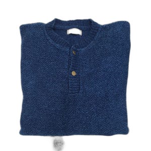 May include: Folded navy blue sweater with a crew neck and a three-button placket. The sweater has a textured knit pattern and gold-colored buttons. The garment is folded neatly, showcasing its shape and design. The sweater is a classic style.