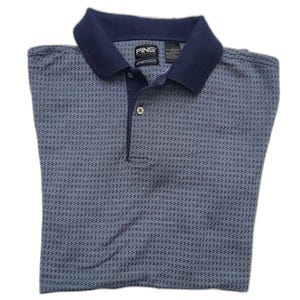May include: A folded blue polo shirt with a navy blue collar and a subtle dotted pattern. The shirt has a three-button placket and the brand name "PING" is visible on the collar.