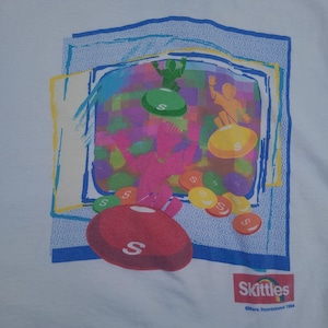 May include: White t-shirt with a vibrant Skittles graphic. The design showcases a large red Skittle, smaller candies, and figures atop the sweets. The Skittles logo and the year 1994 are printed at the bottom of the shirt.