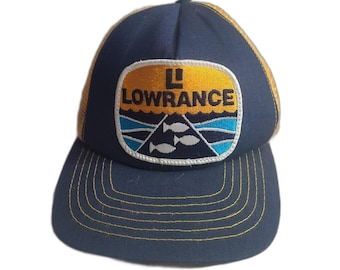 May include: Navy blue trucker hat with a yellow mesh back and a curved brim. The front features a patch with the word "LOWRANCE" in white, a logo, and a design of fish and waves.