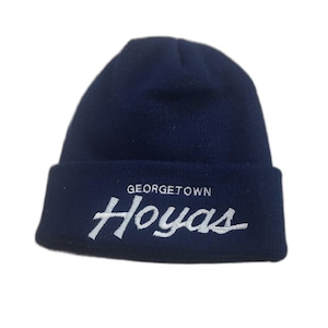 May include: A navy blue knit beanie with the text "GEORGETOWN Hoyas" embroidered in white. The hat has a folded brim and a classic, casual style. Perfect for cold weather.