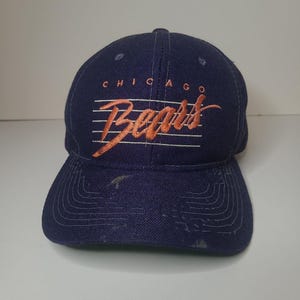 May include: A navy blue baseball cap featuring the word CHICAGO in orange block letters above the word Bears in orange script, with thin horizontal white lines behind the text and visible wear on the brim and crown of the fabric hat.