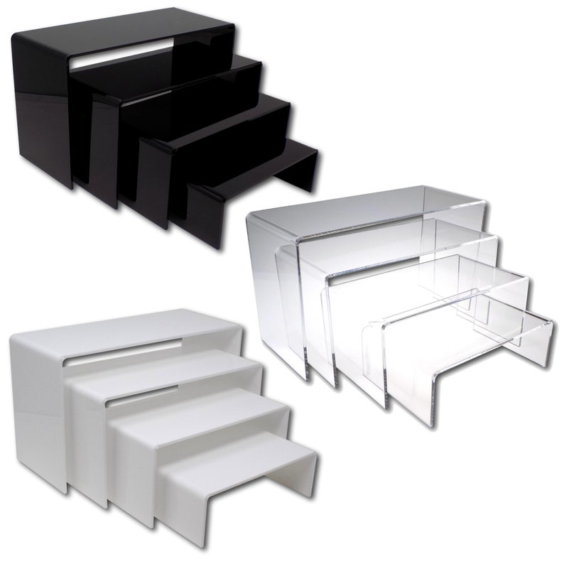 May include: Set of three acrylic display risers in black, clear, and white. The risers are tiered, with the largest at the bottom and the smallest at the top. Each riser has a curved front edge.
