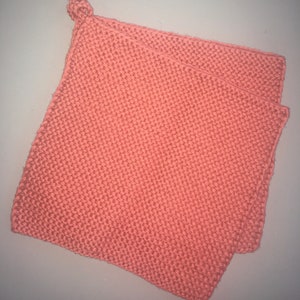 Könnte beinhalten: Ein korallenfarbener, quadratischer Topflappen aus Strick mit einer Schlaufe zum Aufhängen. Der Topflappen ist aus einem dicken, strukturierten Garn gefertigt, und die Maschen sind deutlich sichtbar. Das gefaltete Design bietet zusätzlichen Hitzeschutz.