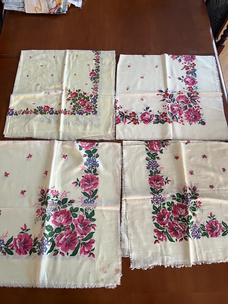 May include: Four white cotton handkerchiefs with a floral pattern in shades of pink, green, and purple. The handkerchiefs have a scalloped edge.