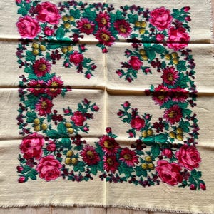 Puede incluir: Un textil cuadrado de color crema con un diseño floral. El diseño presenta grandes rosas rosas, flores rojas más pequeñas, hojas verdes y bayas amarillas dispuestas en un patrón de borde. Los bordes del textil están deshilachados.