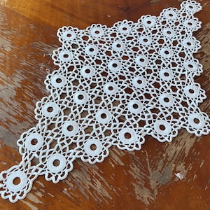 Op de afbeelding: Een witte gehaakte tafelloper met een delicaat patroon van cirkels en bloemen. De loper ligt op een houten tafel.