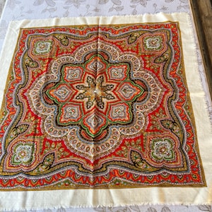 Puede incluir: Un pañuelo cuadrado con un vibrante estampado floral inspirado en el cachemir en tonos rojo, verde y dorado, con un motivo de estrella central. El diseño está enmarcado por un borde ancho de color crema con flecos y detalles simétricos.